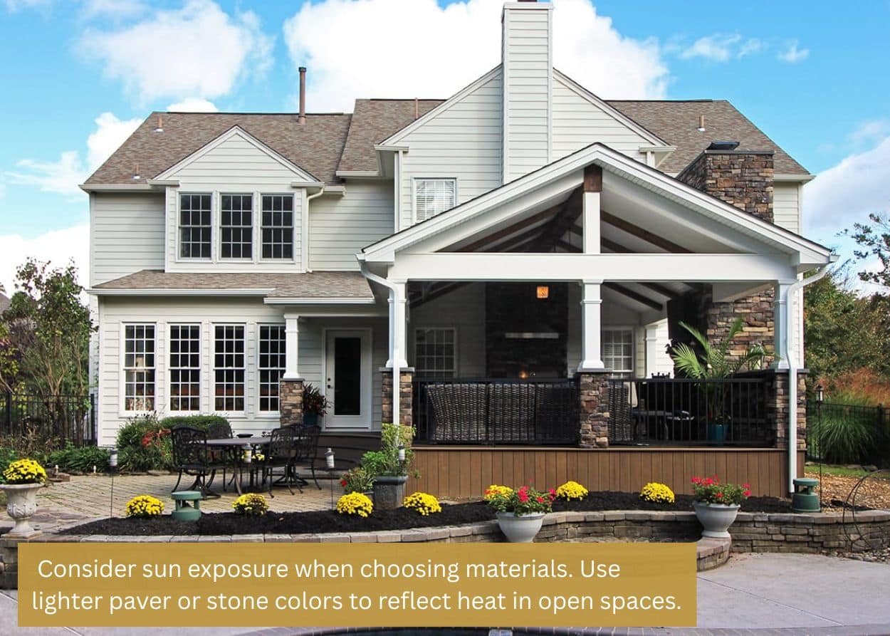 Backyard patio with light-colored pavers, landscaping, outdoor dining set, and a covered porch attached to a two-story home. Caption: Consider sun exposure when choosing materials. Use lighter paver or stone colors to reflect heat in open spaces.
